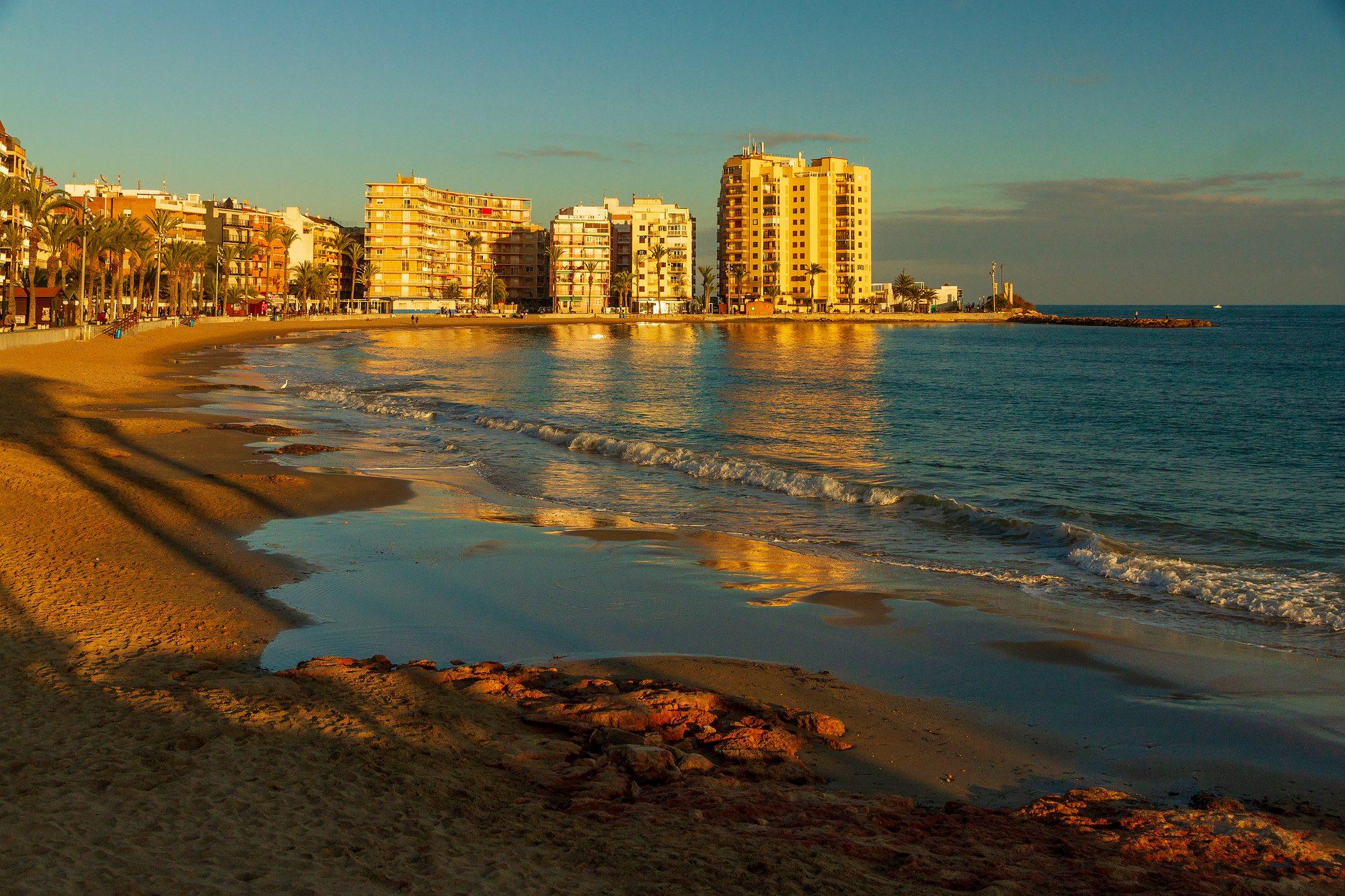 Playa del Cura in Torrevieja in low golden light: a curve of sand with reddish rocks in the foreground, small waves breaking along the shore, and palm trees and apartment blocks glowing in the sun around the bay, which ends in a short breakwater