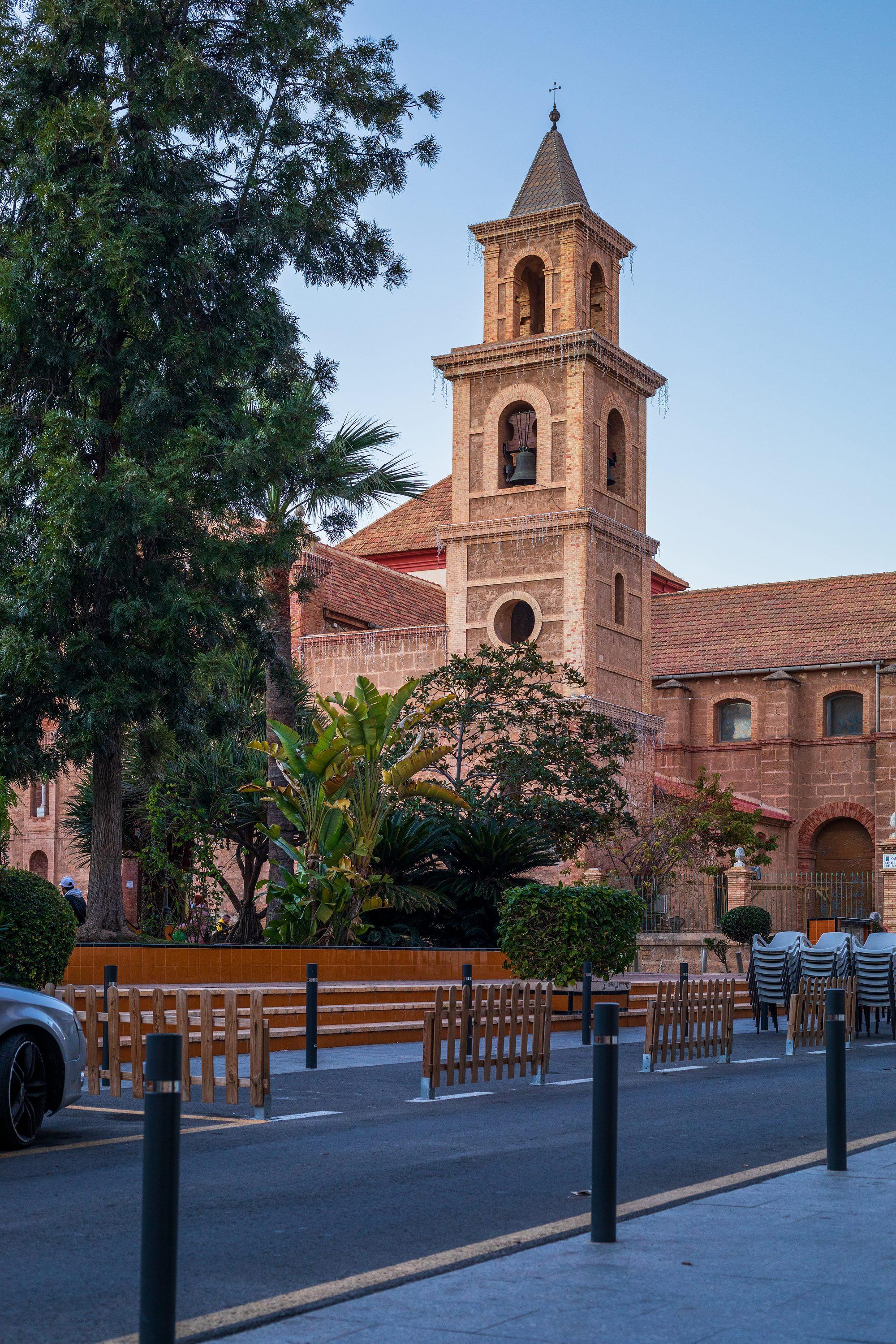 The brick and sandstone bell tower of the Iglesia Arciprestal de la Inmaculada Concepción in Torrevieja, with arched bell openings and a tiled spire topped by a cross, rising above the tiled roofs of the church and the trees, palms and banana plants of the Plaza de la Constitución, with bollards, low wooden fences and stacked café chairs along a quiet street in the foreground