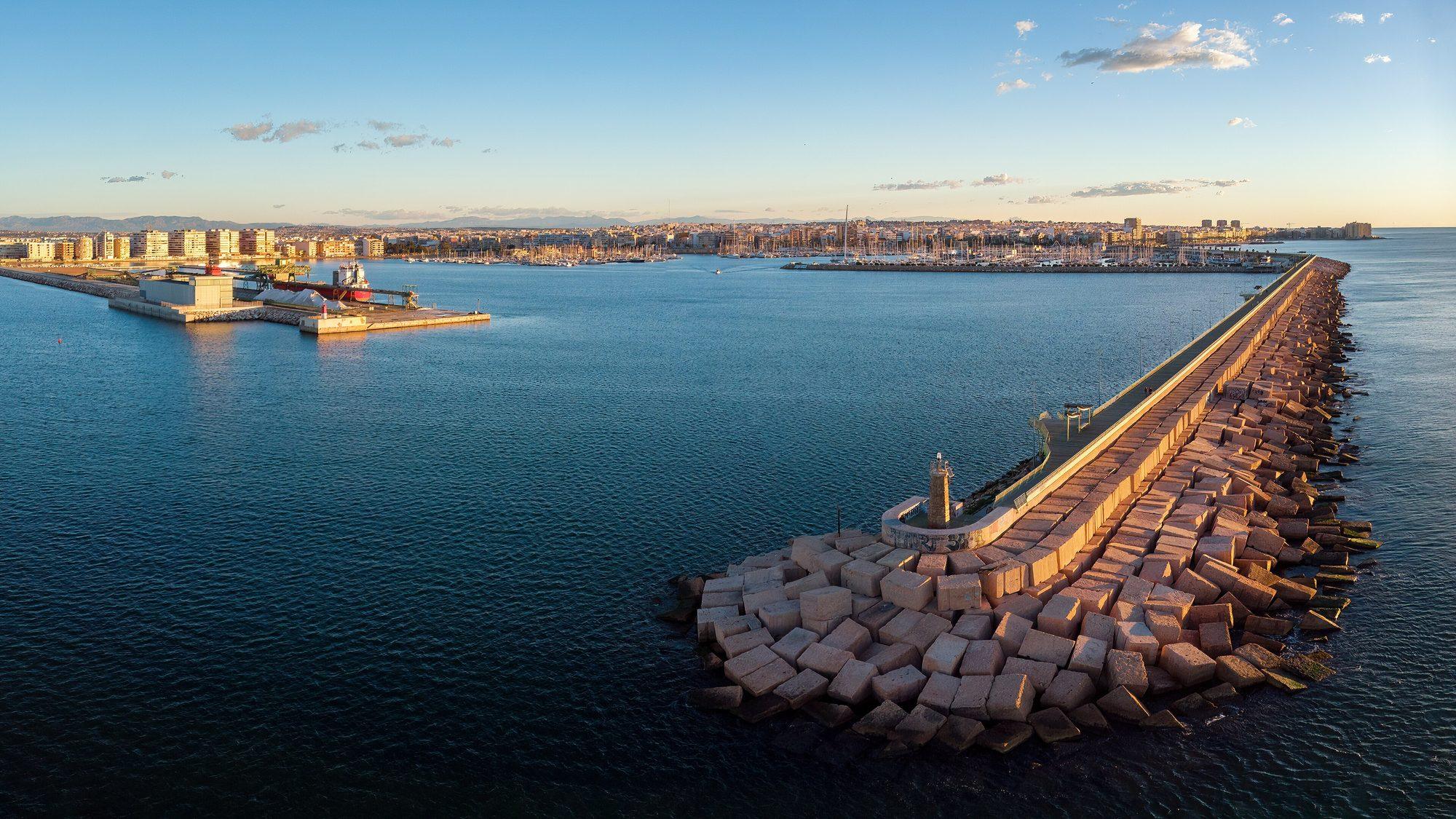 The port of Torrevieja in low morning light, seen from above the sea: the Dique de Levante breakwater, armoured with pale concrete blocks and topped by a promenade, curving out to a small beacon tower at its tip; across the calm blue water lie the marina, the salt-loading quay with a moored cargo ship, and the town's apartment blocks along the shore